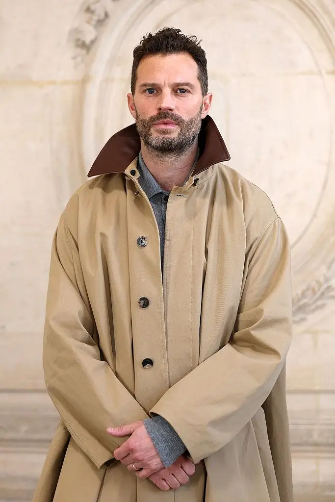 Portrait of Jamie Dornan wearing a beige lightweight coat with a structured collar layered over a knit top, standing against an architectural stone backdrop, conveying a refined, understated approach to transitional outerwear