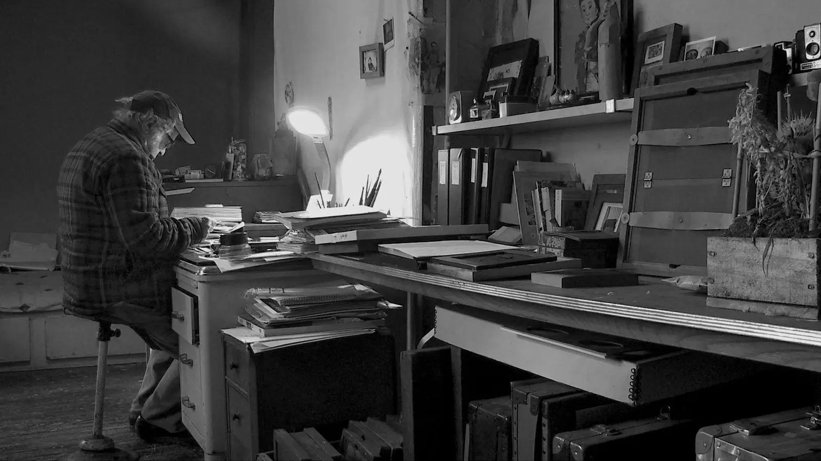 Black-and-white image of Robert Frank seated at a cluttered studio desk, reviewing prints under a lamp, surrounded by books, frames, and archival materials
