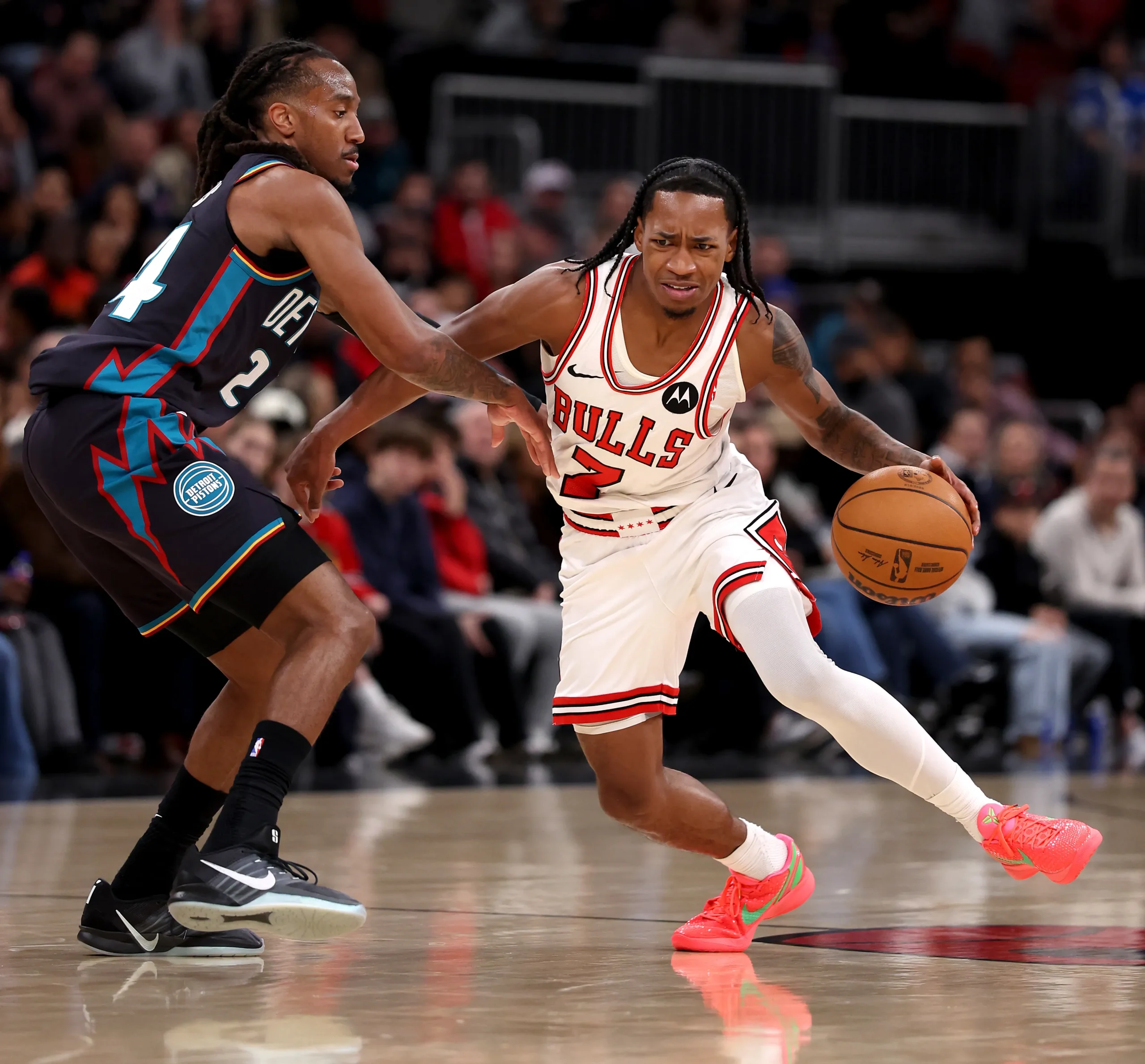 Action shot of a Chicago Bulls guard Rob Dillingham wearing a white #2 jersey driving to the basket while dribbling with his right hand, defended closely by a Detroit Pistons player in a dark uniform, inside a packed arena with blurred crowd in the background