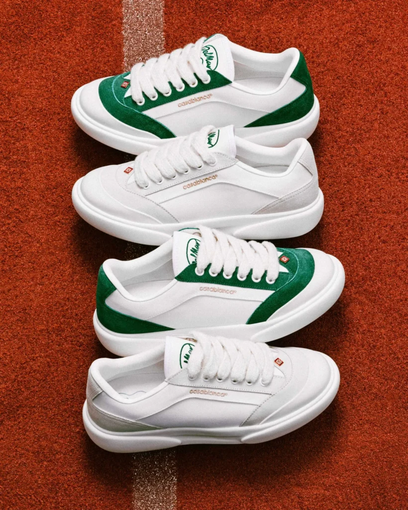 Multiple pairs of Casablanca Del Mar shoe arranged on a clay tennis court, featuring white uppers with green suede accents, white laces, and low-top silhouettes aligned along a court line