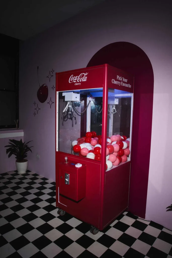 A retro-styled Coca-Cola Cherry claw machine stands as a bold focal point within a softly lit, pastel interior. Finished in vivid cherry red, the machine is filled with glossy red, pink, and white balls, while its glass panels reveal the suspended claw mechanism above. Subtle branding—“Coca-Cola cherry” and “Pick Your Cherry Favourite”—wraps the top edges, reinforcing the theme. The setting balances whimsy and design control: lavender walls with minimal cherry graphics and spark-like accents, a curved red backdrop framing the machine, and a black-and-white checkered floor that nods to classic diner aesthetics