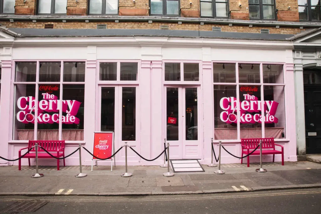 A street-facing view of “The Cherry Coke Café” presents a pastel-pink storefront that merges classic British architecture with bold, contemporary branding. Large window graphics in vivid pink and white announce the concept, with “The Cherry Coke Café” lettering layered over playful cherry motifs. The symmetrical façade features tall glass panes and a central door, framed by clean white trim. Outside, bright pink benches sit on either side, while chrome stanchions and black ropes create a controlled, event-style entrance along the pavement. A small signboard reinforces the branding at street level. The overall scene feels curated and highly photogenic—less a traditional café, more a temporary immersive installation designed to draw attention and invite interaction from passersby