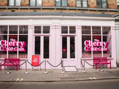 A street-facing view of “The Cherry Coke Café” presents a pastel-pink storefront that merges classic British architecture with bold, contemporary branding. Large window graphics in vivid pink and white announce the concept, with “The Cherry Coke Café” lettering layered over playful cherry motifs. The symmetrical façade features tall glass panes and a central door, framed by clean white trim. Outside, bright pink benches sit on either side, while chrome stanchions and black ropes create a controlled, event-style entrance along the pavement. A small signboard reinforces the branding at street level. The overall scene feels curated and highly photogenic—less a traditional café, more a temporary immersive installation designed to draw attention and invite interaction from passersby