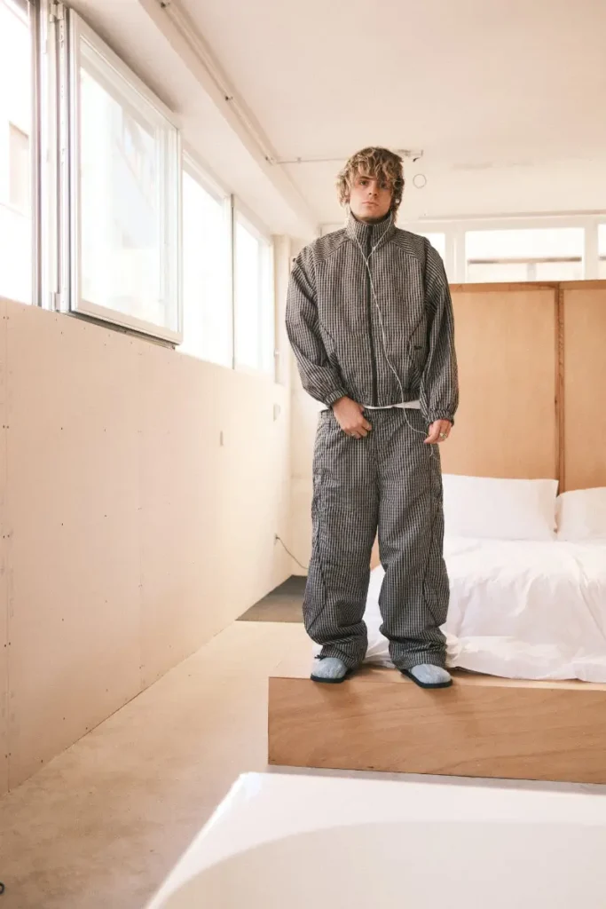 A person stands on a low wooden platform in a softly lit, minimalist room, wearing a matching textured jacket and loose trousers in a muted gray pattern, paired with light-colored slip-on shoes. Natural light filters through large windows along the wall, while a simple bed with white bedding and a wooden partition sits behind them, reinforcing the calm, pared-back interior aesthetic