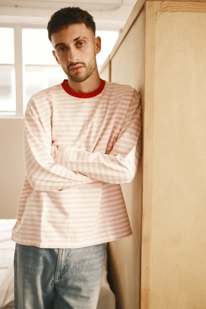 A person leans against a light wooden partition in a softly lit room, wearing a pink-and-white striped long-sleeve shirt with a red collar and relaxed blue jeans. Natural light filters through the windows behind, highlighting their neutral expression and casual posture, while the minimal interior and warm tones create a calm, understated atmosphere