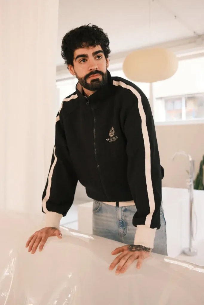 A person leans forward with both hands resting on the edge of a white bathtub in a bright, minimalist interior, wearing a black zip-up jacket with white stripe detailing along the sleeves and light blue jeans. Soft natural light fills the space, highlighting their thoughtful expression and the clean, modern setting with neutral tones and simple furnishings