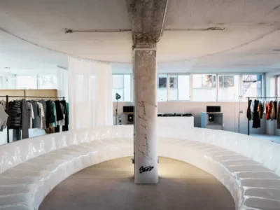 A minimalist studio space features a large, curved white seating arrangement encircling a central column with subtle markings. Clothing racks line the perimeter, displaying neatly arranged garments, while speakers and equipment sit near the windows. Soft daylight filters through the room, creating a clean, gallery-like setting that feels prepared for a presentation or showroom event