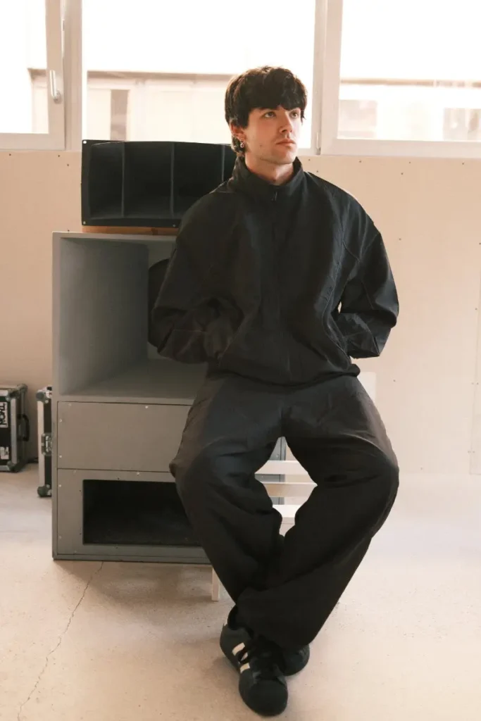 A person sits on a low stool in a bright, minimalist studio, dressed in an all-black outfit with a zip-up jacket and loose trousers paired with dark sneakers. Positioned in front of a large speaker cabinet, they hold a composed, slightly turned gaze, while soft daylight from the windows creates a clean, understated atmosphere around the scene