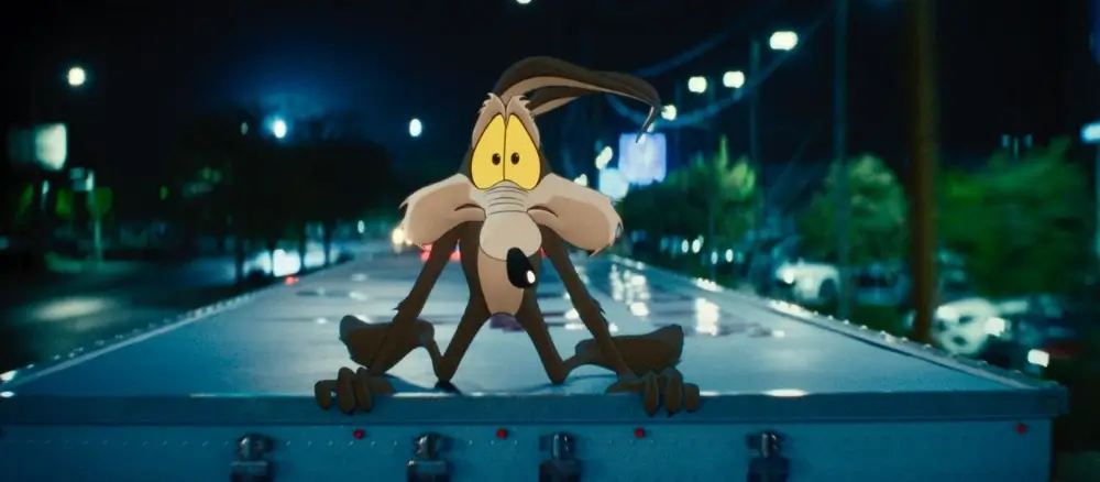 Wile E. Coyote crouched on the back of a moving truck at night in a live-action city street, wide-eyed under streetlights in a scene from Coyote vs. Acme