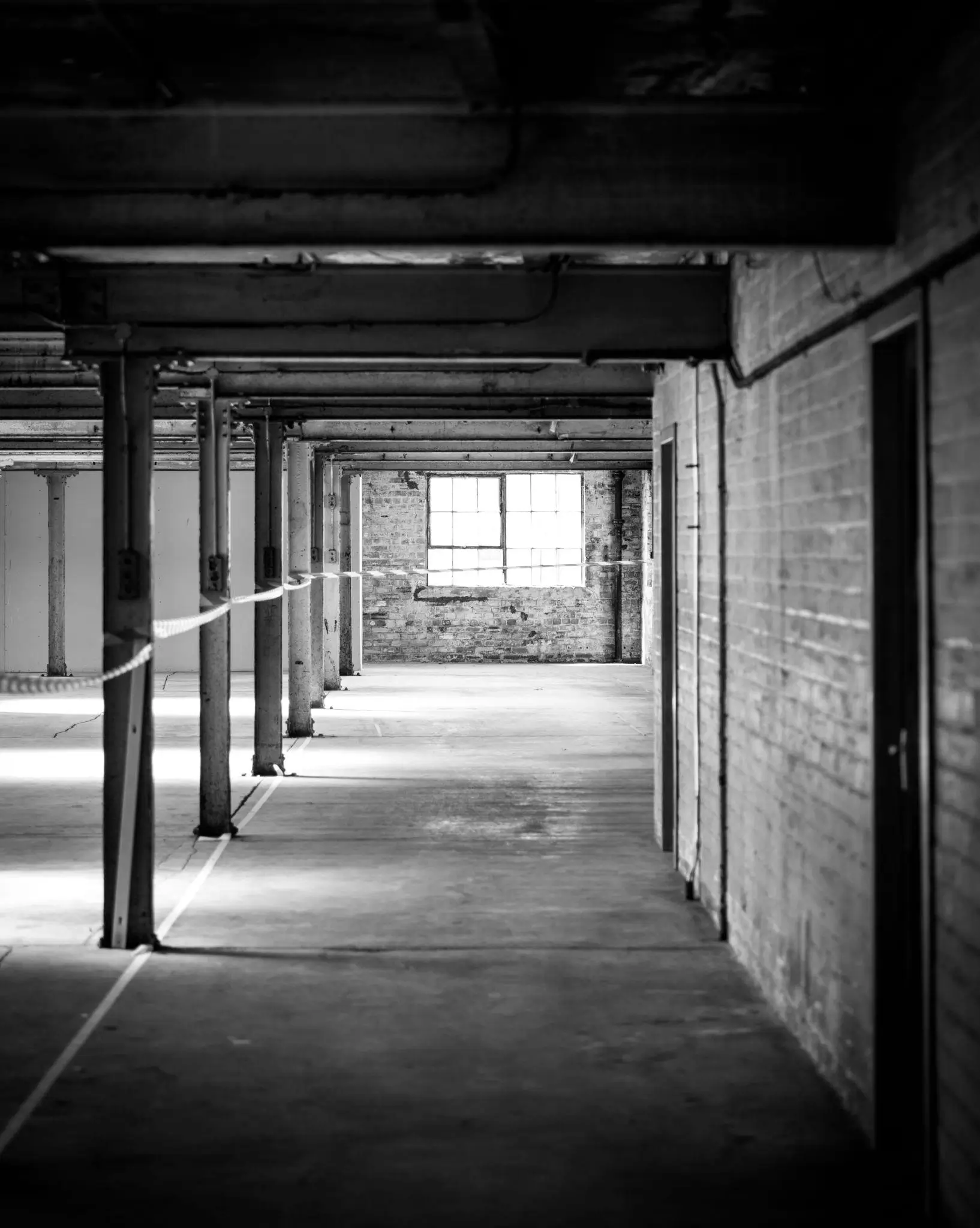 A black-and-white view of an empty industrial interior stretches into the distance, defined by exposed brick walls, a row of evenly spaced support columns, and overhead beams. Light pours in from a window at the far end, creating strong contrast and long shadows across the concrete floor, emphasizing the raw, architectural structure and quiet, atmospheric depth of the space