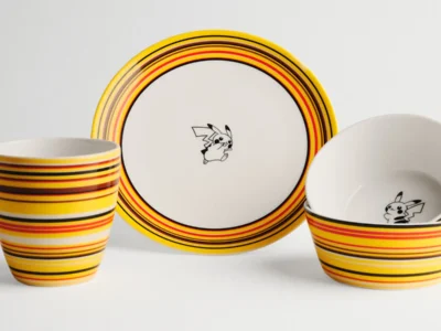 A coordinated ceramic tableware set featuring a plate, mug, and two bowls, all designed with bold horizontal stripes in yellow, orange, red, and black. Each piece includes a small black-and-white illustration of Pikachu placed centrally inside the plate and bowls. The items are arranged against a clean white background, highlighting the vibrant color bands and play character detail