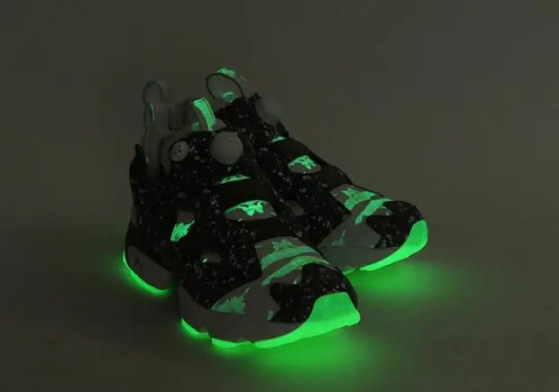 A pair of futuristic shoe glow vividly in the dark, featuring neon green illumination along the outsole and toe. The upper is predominantly black with scattered camo-like patterns and speckled detailing, while translucent elements and structural overlays hint at a tech-driven design. The shoes are photographed against a dim background, emphasizing the luminous green glow and cyberpunk-inspired aesthetic