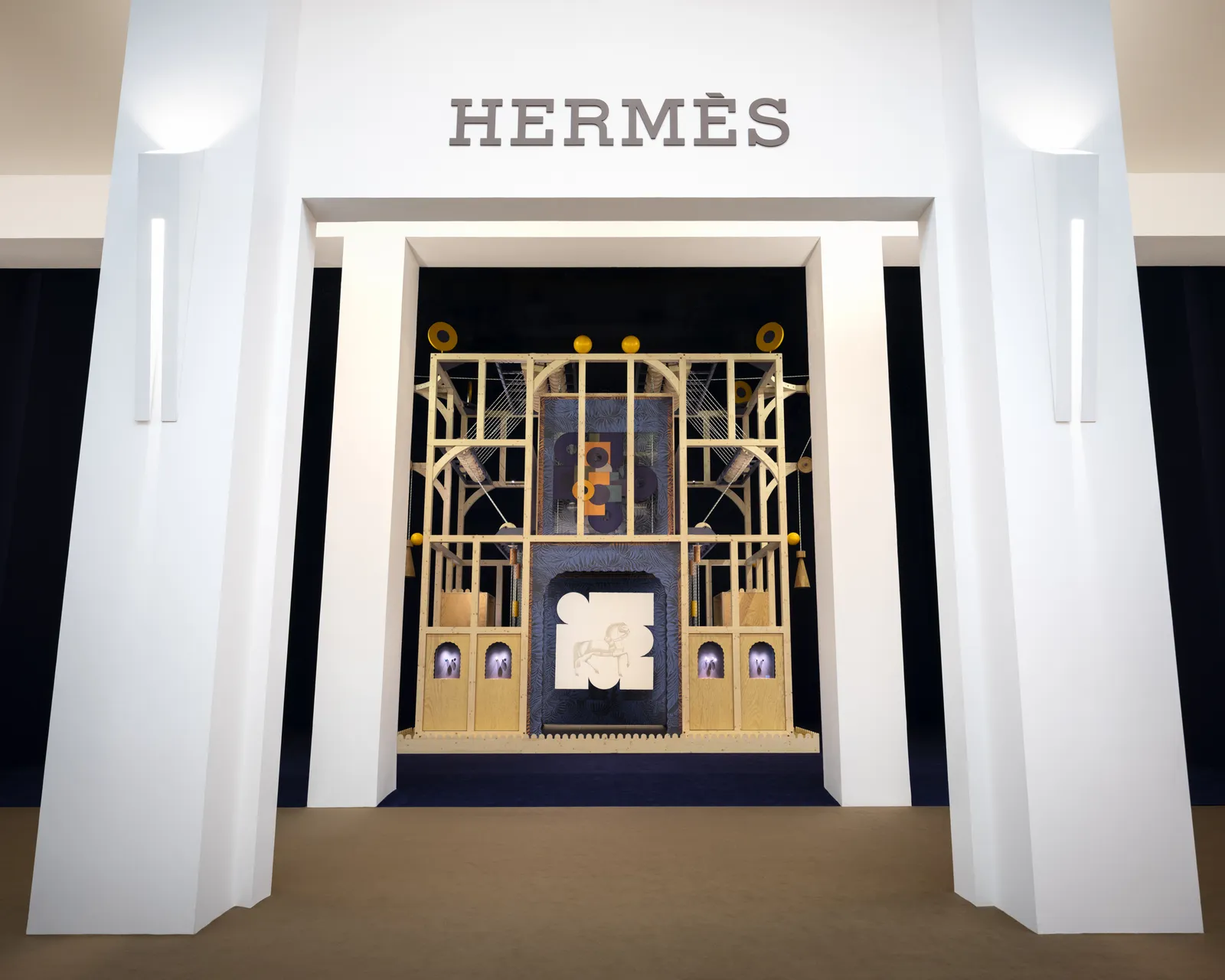 Minimalist Hermès pavilion entrance with a white architectural frame and the “HERMÈS” logo above, revealing an ornate interior installation composed of a symmetrical wooden structure with kinetic, clockwork-like elements and softly lit display niches
