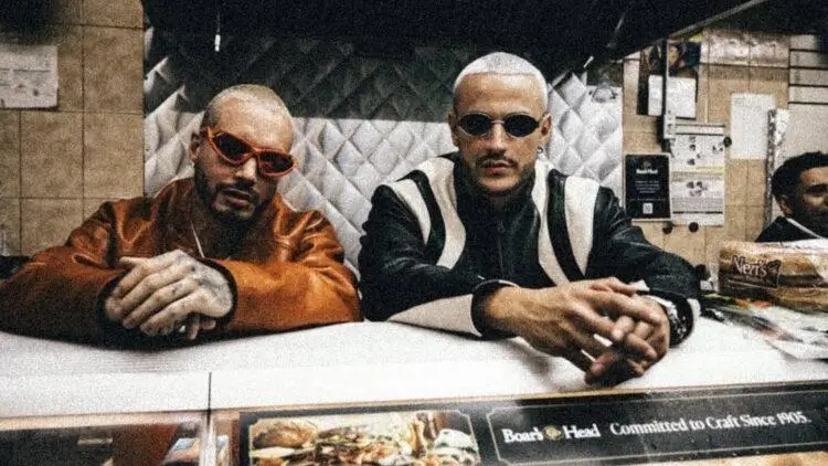 J Balvin left side and right side DJ Snake behind deli-style counter, staring directly at the camera with a composed, assertive presence. Both wear dark sunglasses and have closely shaved or bleached hair, giving a stark, stylized contrast. One is dressed in a satin-like burnt orange jacket, while the other wears a black-and-white leather jacket with bold paneling. Behind them, the setting appears to be a modest food shop kitchen with tiled walls and stainless steel surfaces, while the foreground shows part of a menu display. The image carries a gritty, cinematic feel—part street portrait, part fashion editorial