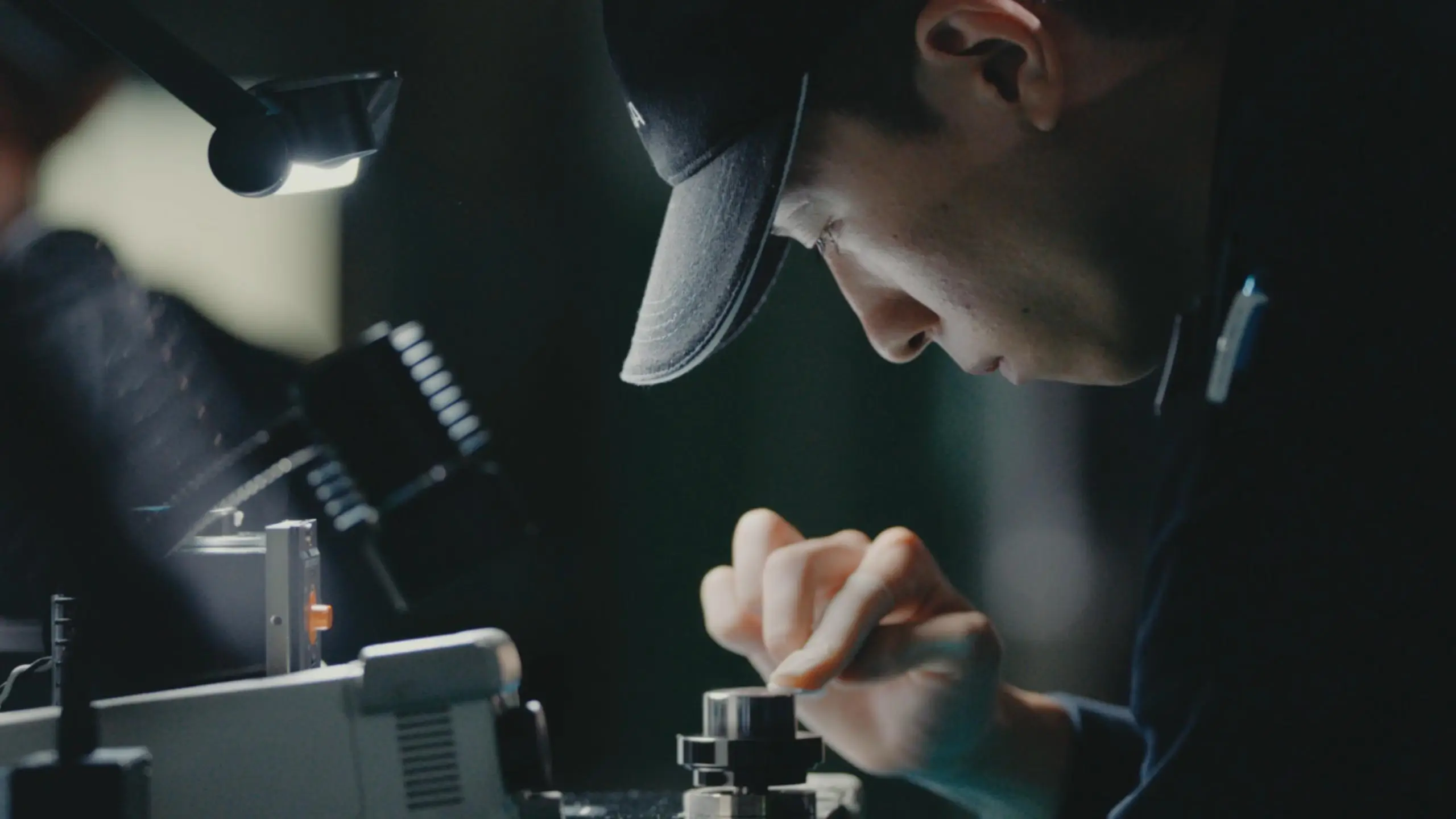 A technician wearing a cap carefully adjusts a small camera component under a focused work light, highlighting precision craftsmanship in a dimly lit workspace