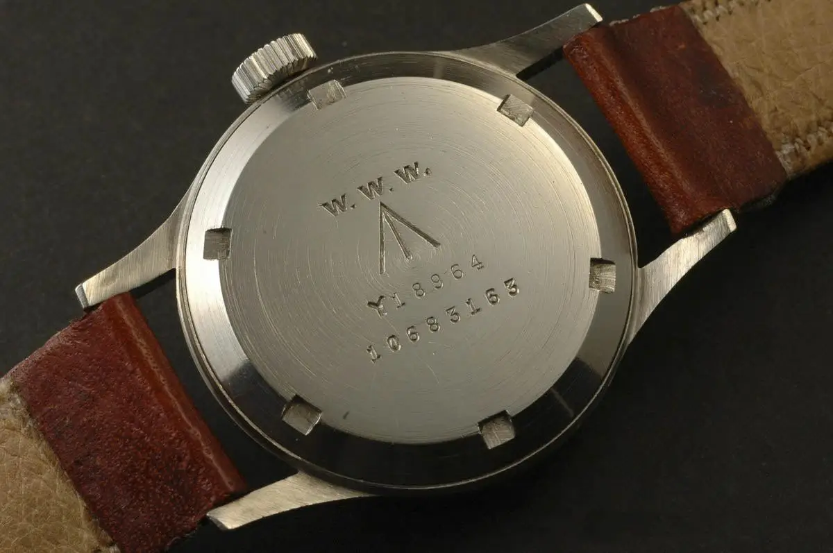 The brushed stainless steel caseback of a vintage military-issued wristwatch is shown in close detail, engraved with “W.W.W.” (Wrist, Watch, Waterproof), a broad arrow military mark, and serial numbers. The watch is fitted with a worn brown leather strap, its patina and utilitarian markings emphasizing its historical, service-driven origin