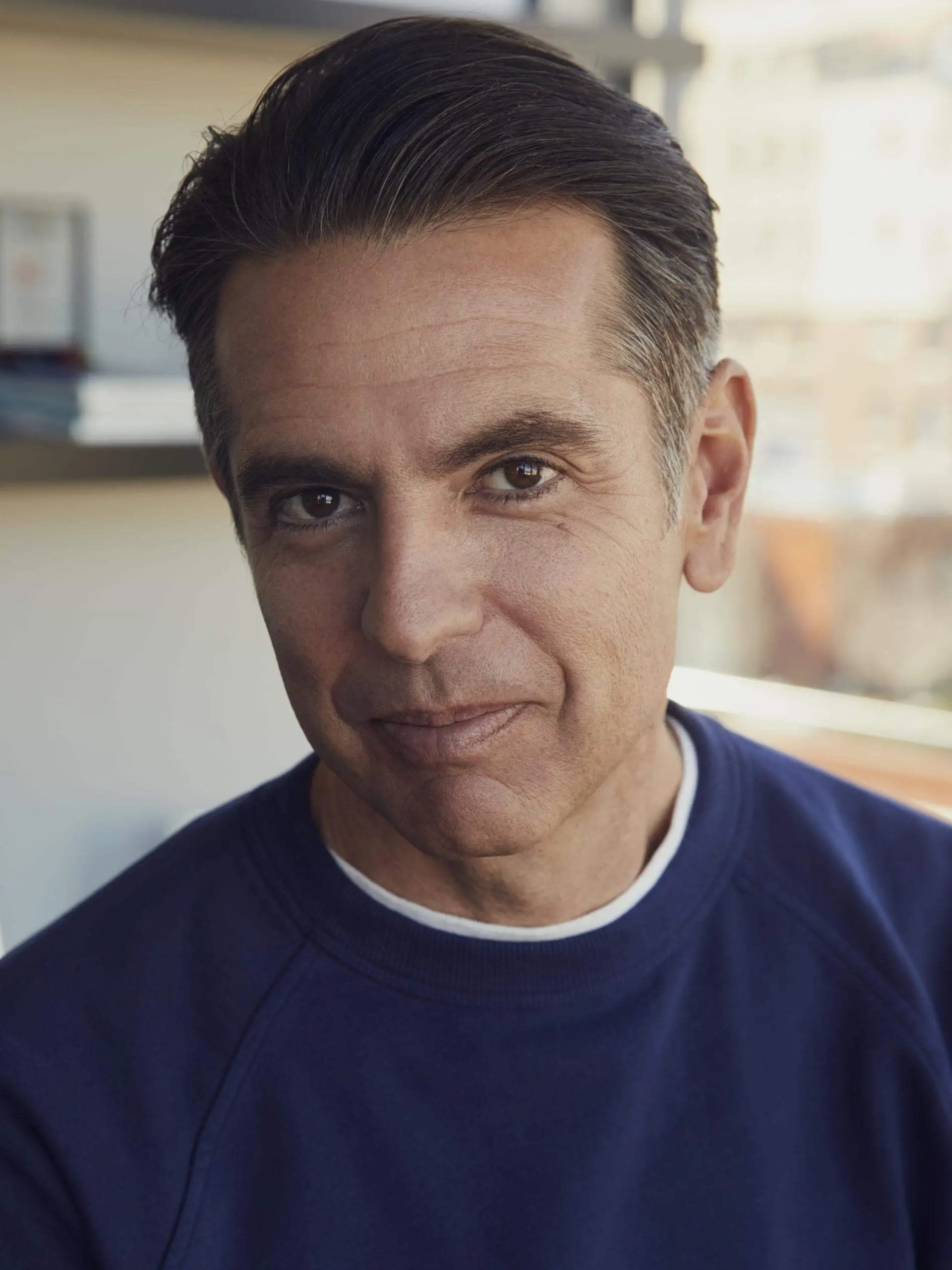 Close-up portrait of Matt Pincus with neatly combed dark hair and a slight, relaxed smile, looking directly at the camera. He wears a navy crewneck sweatshirt, and soft natural light highlights his face, bringing out subtle skin texture and warm brown eyes. The background is softly blurred, suggesting an indoor setting with shelves and a window, giving the image a calm, approachable, and contemporary feel