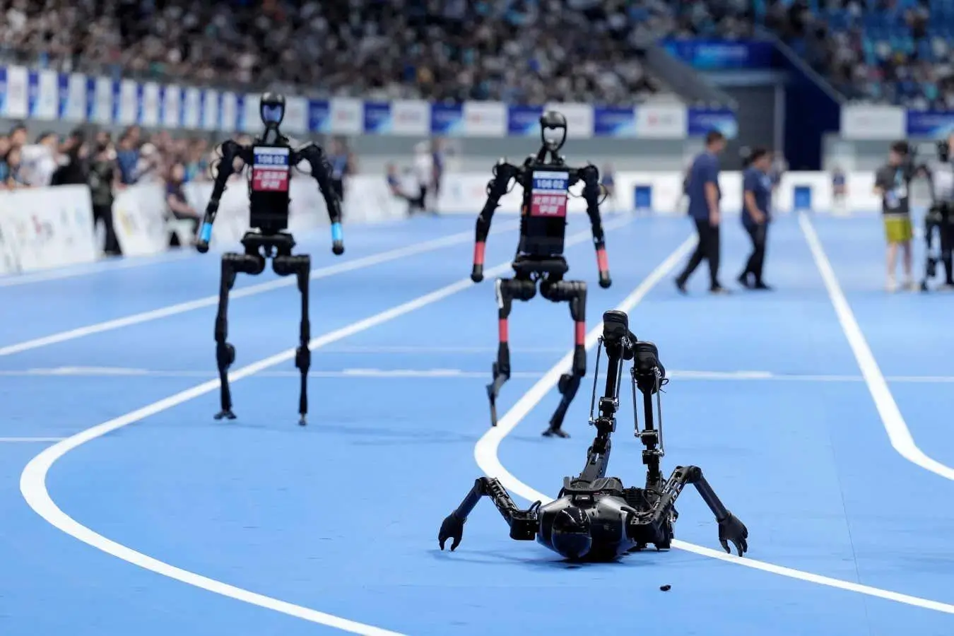 Humanoid robots race on an indoor track as one robot falls onto its back in the foreground while others continue running during a robotics competition