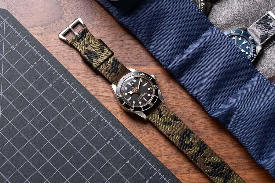 A stainless steel dive watch with a black dial and rotating bezel rests on a wooden surface, fitted with a green camouflage fabric strap. Nearby, a precision cutting mat and a padded blue watch roll partially reveal another timepiece, creating a utilitarian, workshop-inspired setting that emphasizes durability and field-ready design