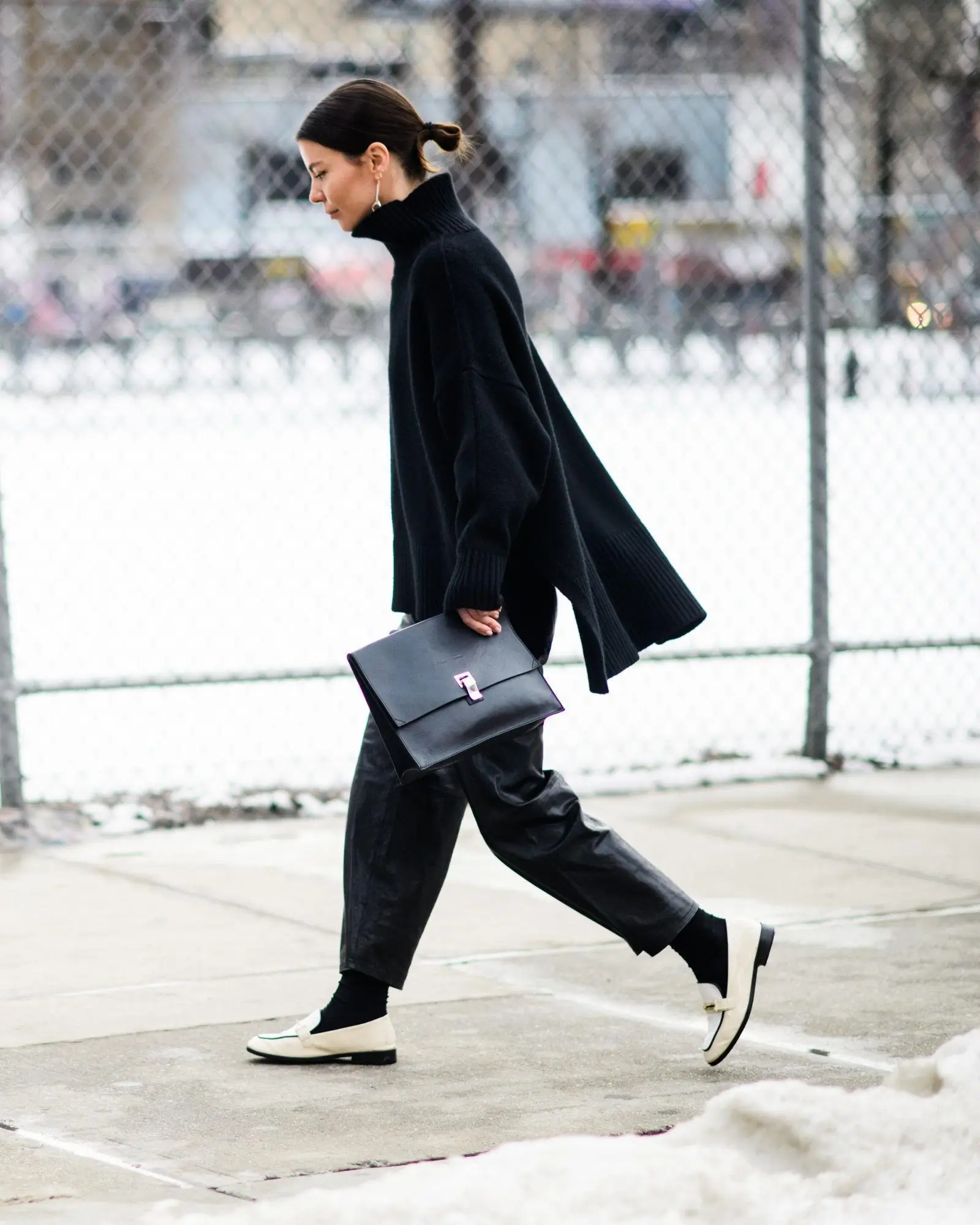 Chic winter street style: woman in oversized black turtleneck sweater, leather pants, and white loafers walking in the snow with a black leather bag. Clean minimalist look
