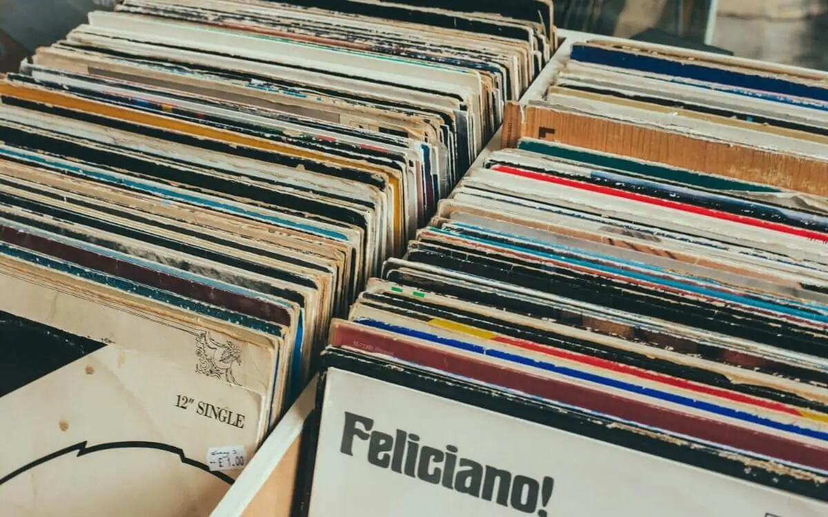 A close-up view of wooden crates filled with vintage vinyl records, their worn sleeves tightly packed and slightly frayed at the edges. The records are organized upright in rows, revealing a mix of aged covers and labels, including a visible divider marked “12" SINGLE” and a partially visible album sleeve reading “Feliciano!” The scene evokes the tactile, analog charm of record digging in a secondhand shop or market