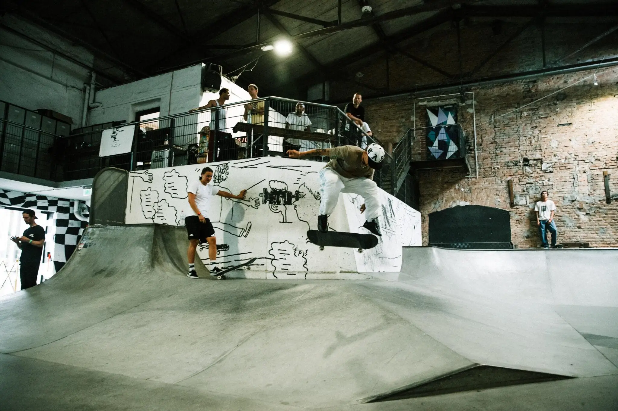 Skater mid-trick in a raw industrial indoor skatepark, launching off a graffiti-covered ramp with onlookers watching. Pure skate session energy