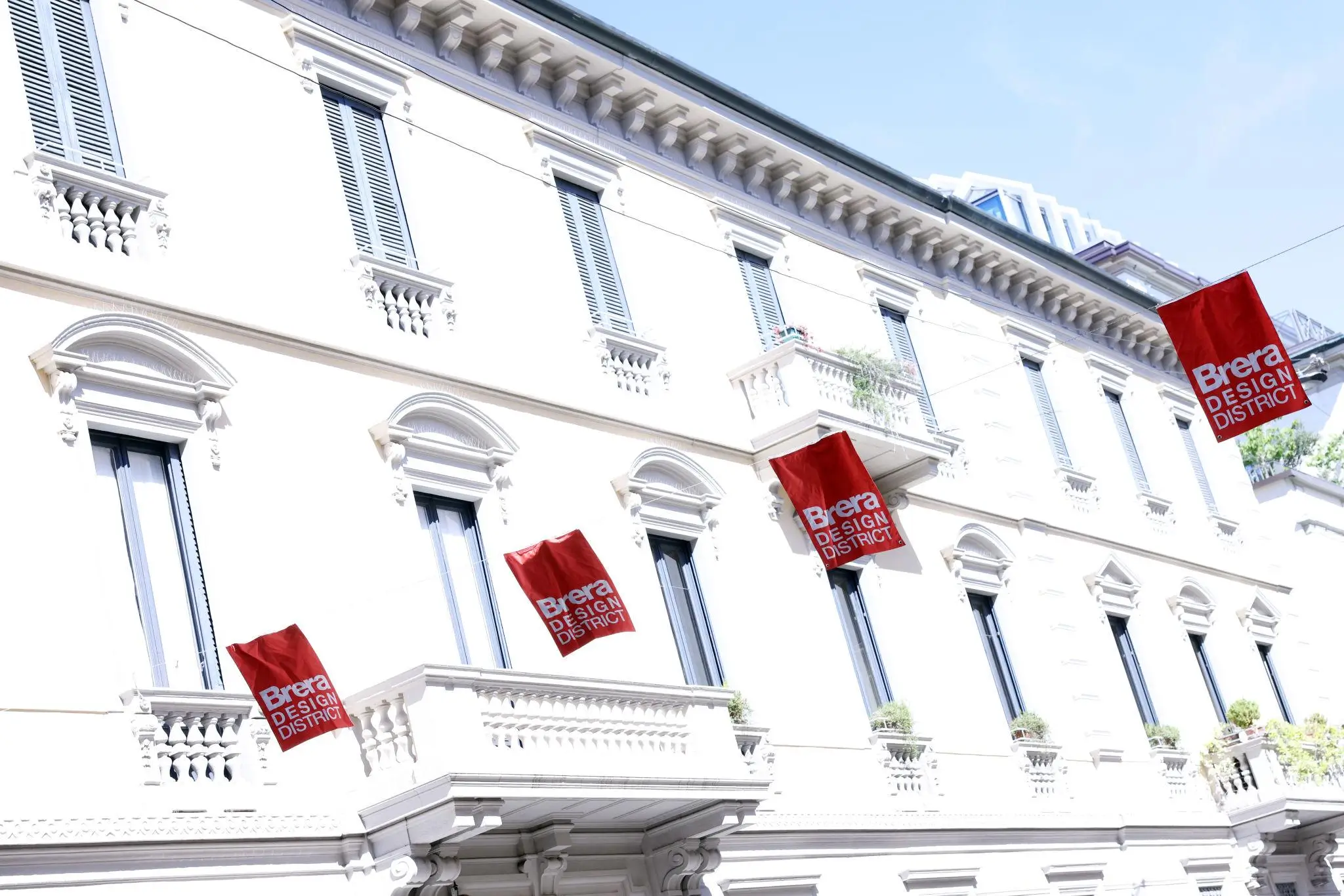 A historic white building facade with ornate balconies and shuttered windows displays multiple red “Brera Design District” banners, set against a bright blue sky