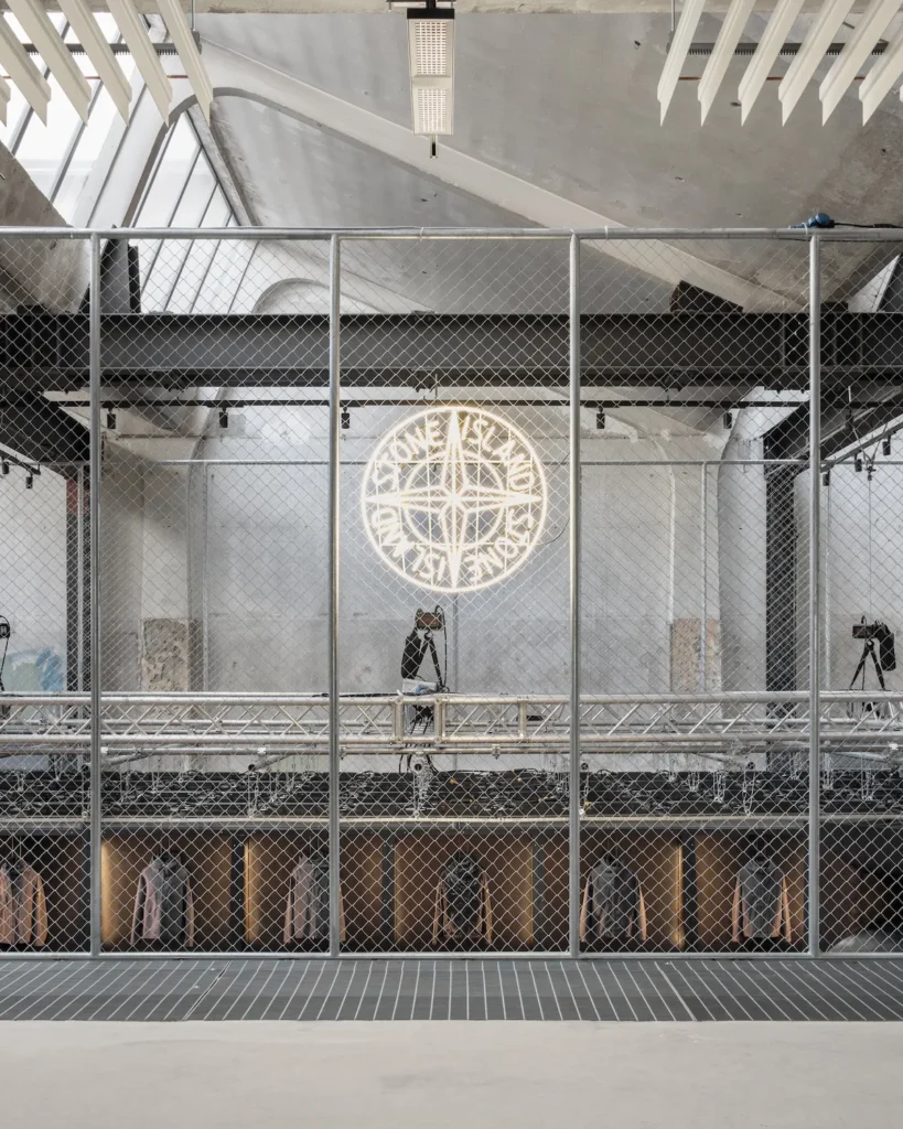 An industrial showroom interior framed by a chain-link mesh barrier, revealing a two-level retail environment beyond. At the center, a glowing circular neon emblem resembling a compass is mounted against a raw concrete wall, casting warm light across the space. Below, a row of recessed display niches presents neatly hung garments, each softly illuminated. Above, an exposed ceiling with angular skylights and suspended acoustic panels enhances the utilitarian aesthetic, while a lone figure stands on the upper level, adding a sense of scale and quiet observation