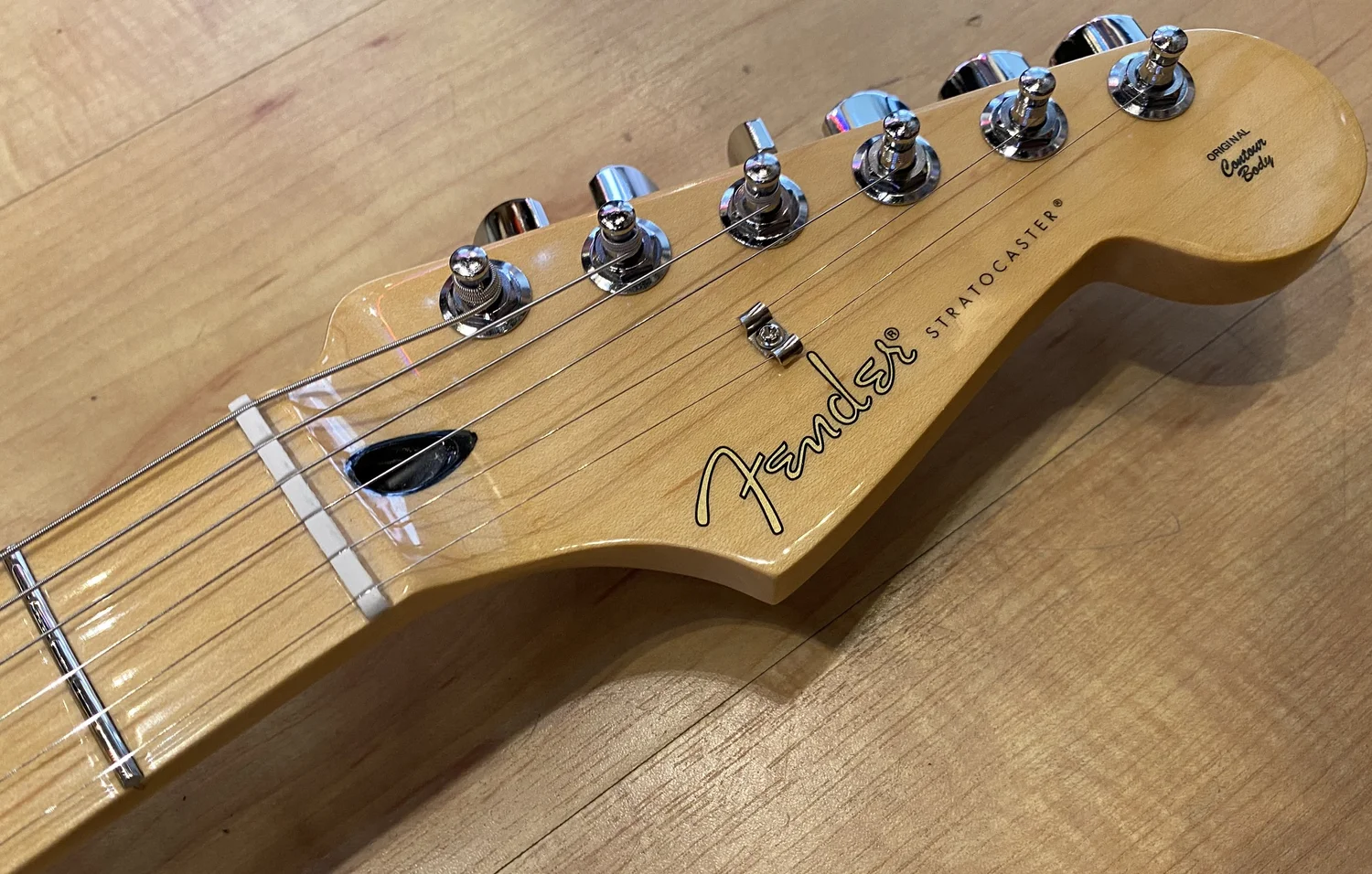 Close-up of Fender Player Stratocaster headstock with maple neck, chrome tuners, Fender logo, and 'Original Contour Body' marking on a wooden surface