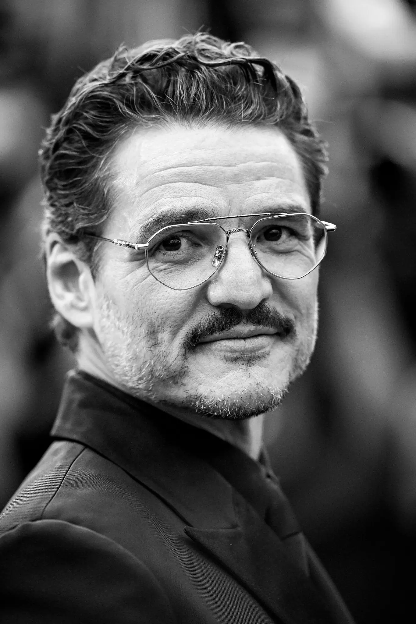 Black-and-white close-up portrait of Pedro Pascal turning slightly toward the camera with a calm, self-assured expression, wearing thin metal aviator-style glasses and a structured dark jacket; soft lighting accentuates the texture of his hair, light stubble, and subtle smile against a blurred, urban background