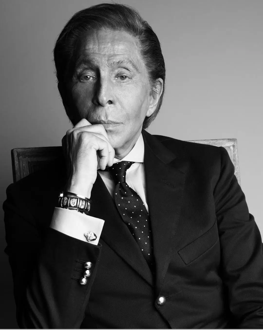 A black-and-white portrait of Valentino Garavani seated in a tailored dark suit and polka-dot tie, resting his chin on his hand; he wears a bracelet and cufflinks, with neatly styled hair and a composed, introspective expression against a plain backdrop