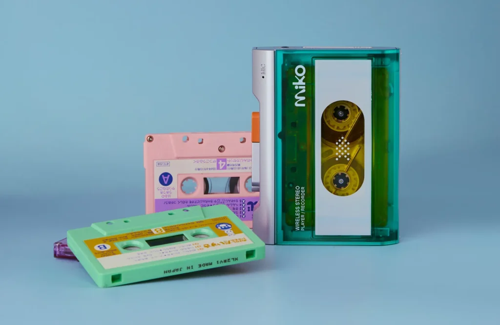 Gadhouse Miko cassette player in transparent green with exposed tape reels, surrounded by colorful cassette tapes on a blue background, highlighting analog music revival and retro audio design