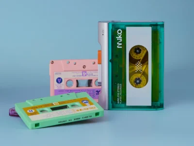 Gadhouse Miko cassette player in transparent green with exposed tape reels, surrounded by colorful cassette tapes on a blue background, highlighting analog music revival and retro audio design