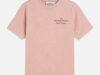 A soft pink short-sleeve T-shirt is displayed flat against a clean, neutral background, showing the front view of the garment. The fabric has a slightly washed, vintage texture, and a small embroidered detail sits on the left chest featuring a crown motif above the text “Jean-Michel Basquiat” and “Scotch & Soda” in a handwritten-style script. The minimalist front contrasts with the more graphic back design, emphasizing subtle branding and a refined, art-inspired aesthetic