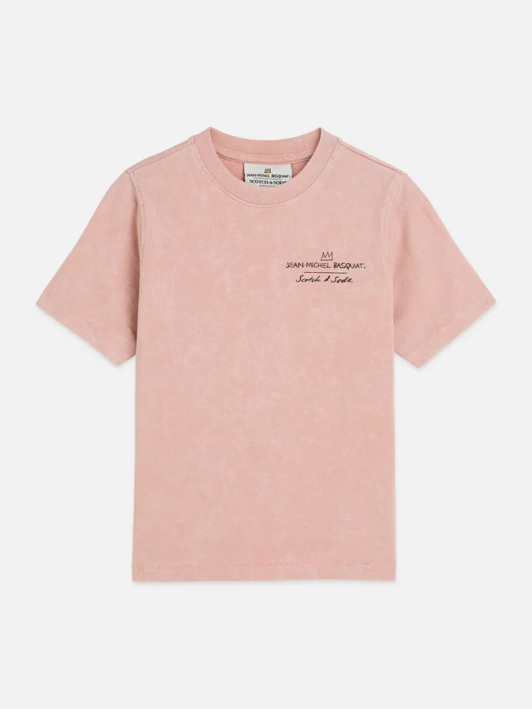 A soft pink short-sleeve T-shirt is displayed flat against a clean, neutral background, showing the front view of the garment. The fabric has a slightly washed, vintage texture, and a small embroidered detail sits on the left chest featuring a crown motif above the text “Jean-Michel Basquiat” and “Scotch & Soda” in a handwritten-style script. The minimalist front contrasts with the more graphic back design, emphasizing subtle branding and a refined, art-inspired aesthetic