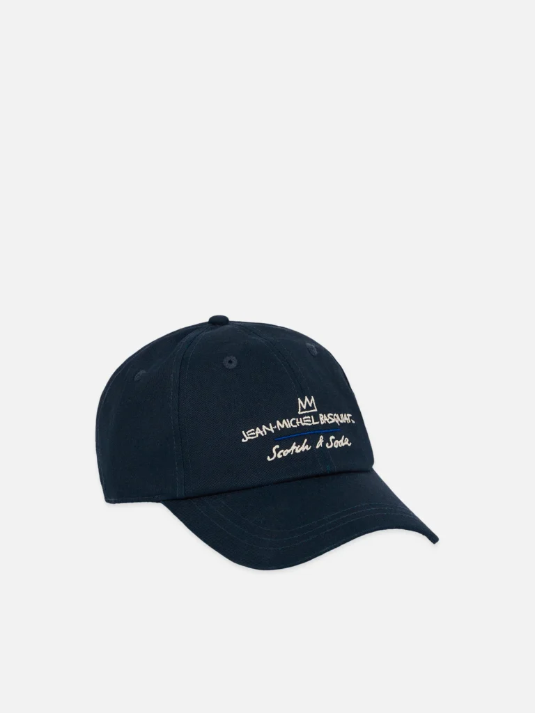 A navy blue baseball cap is displayed against a neutral background, featuring a curved brim and structured crown with stitched ventilation eyelets. Embroidered in white on the front panel is a minimalist crown motif above the text “Jean-Michel Basquiat,” with “Scotch & Soda” written below in a handwritten-style script. The design is clean and understated, blending classic headwear with subtle, art-inspired branding