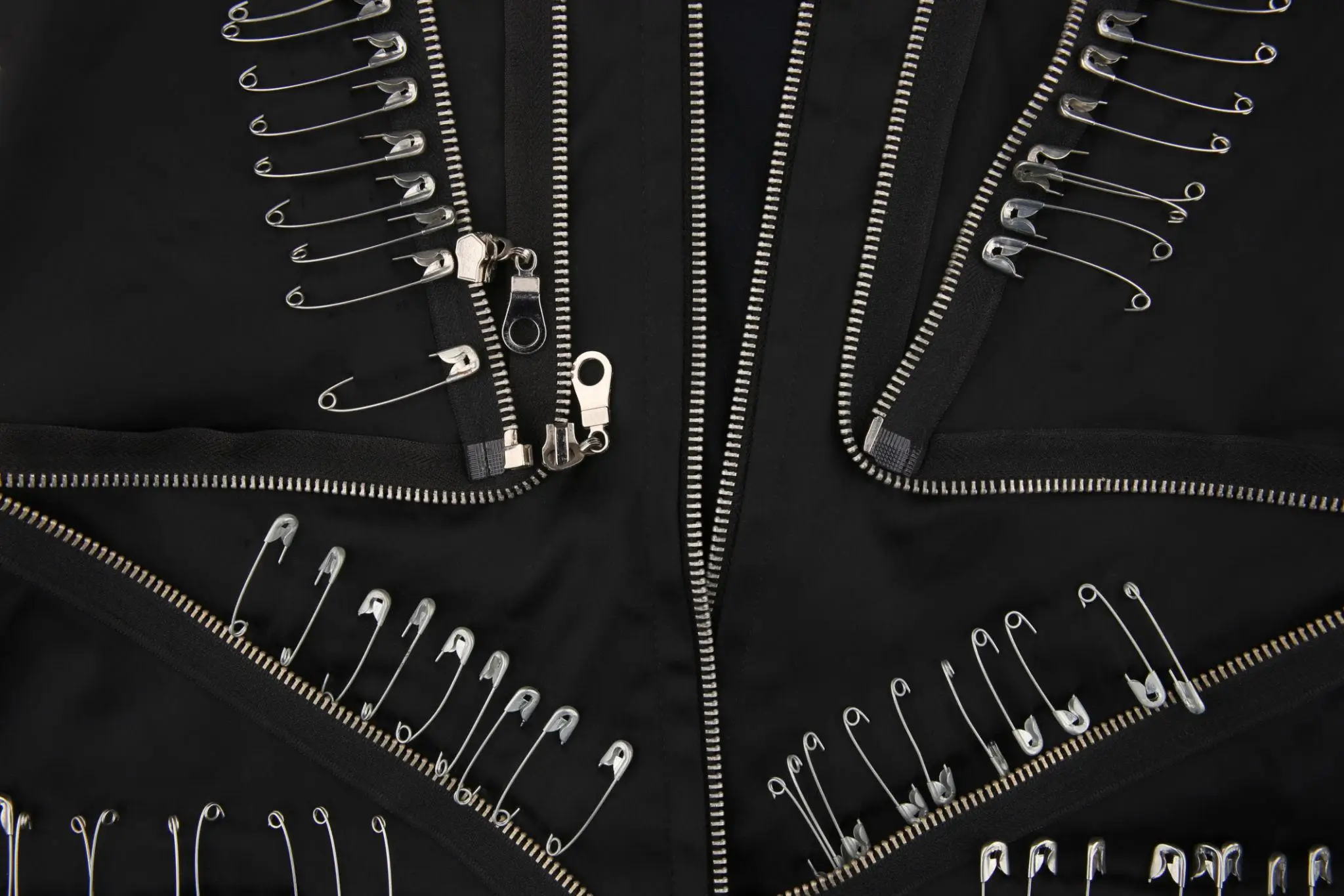 Close-up detail of a black garment emphasizing hardware-heavy construction and deconstructed design. The surface is structured around multiple silver zippers arranged in angular, intersecting lines, creating a geometric framework across the fabric. Along the seams and zipper edges, rows of silver safety pins are meticulously attached, forming linear patterns that echo the garment’s structure while adding a raw, punk-inspired edge. The pins vary slightly in size and orientation, giving a sense of intentional irregularity. The zipper pulls and metal teeth catch the light, contrasting sharply against the matte black fabric. Overall, the composition highlights an industrial, DIY aesthetic—merging utilitarian hardware with rebellious detailing to create a bold, textural statement