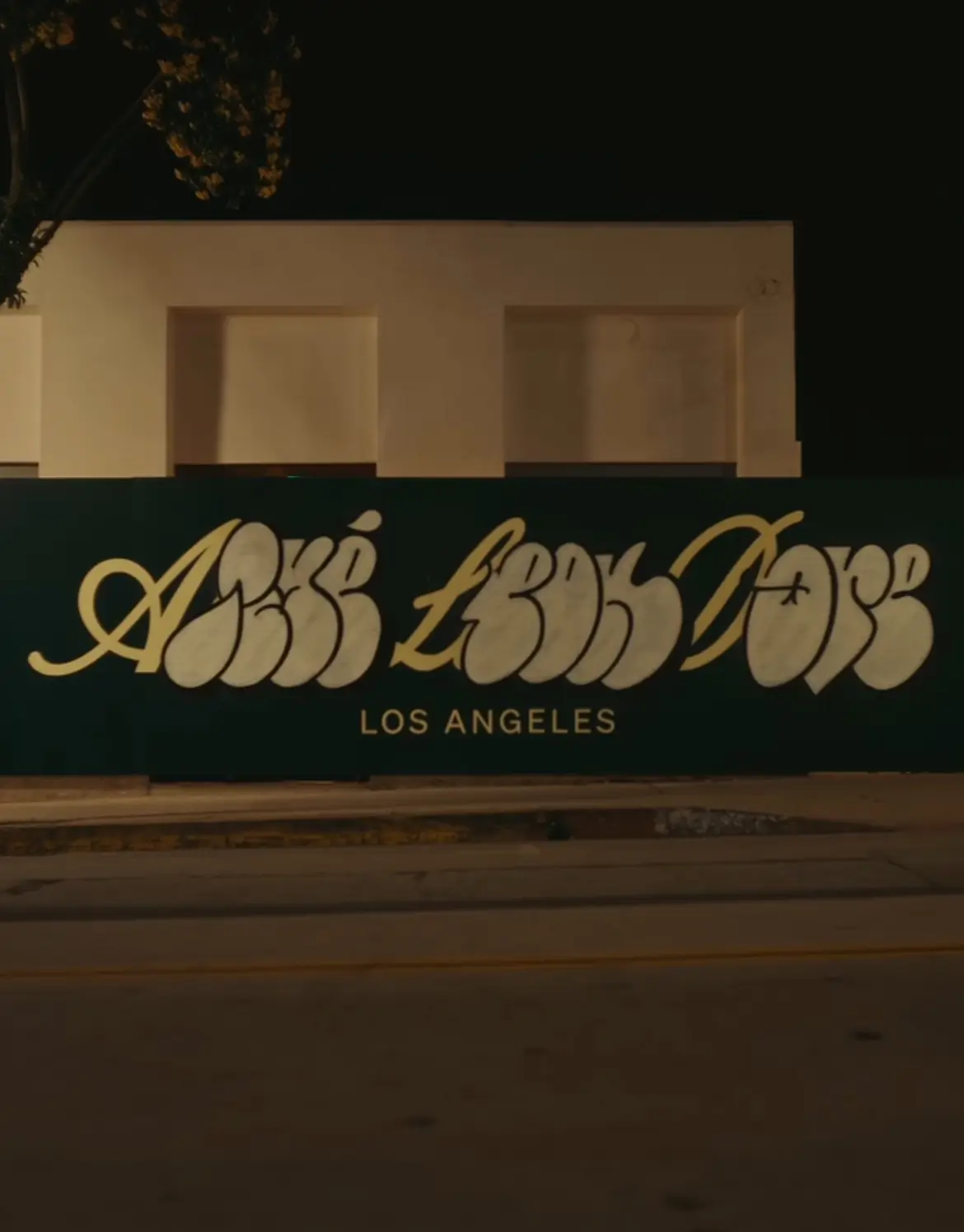 Aimé Leon Dore Los Angeles storefront at night with large cursive logo signage across a dark green facade on Melrose Avenue in West Hollywood, softly lit building and tree above, empty street in foreground