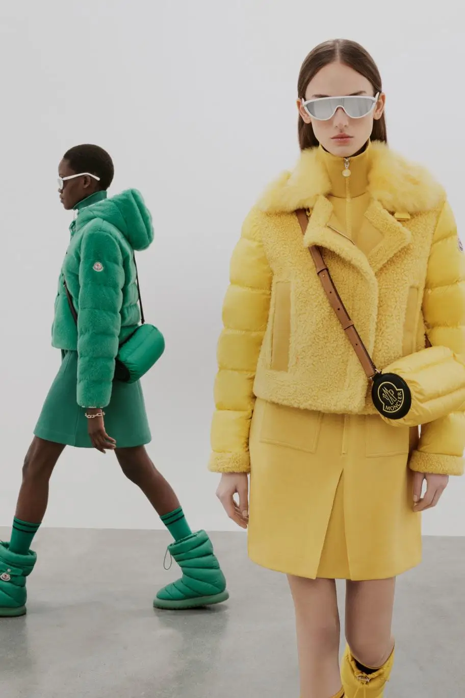 Two models styled in coordinated looks by Moncler, one in a monochrome yellow ensemble featuring a shearling-trimmed puffer jacket, skirt, and crossbody bag with oversized sunglasses, and another in a tonal green outfit with a cropped puffer, skirt, and matching boots, highlighting bold color blocking and sculptural layering
