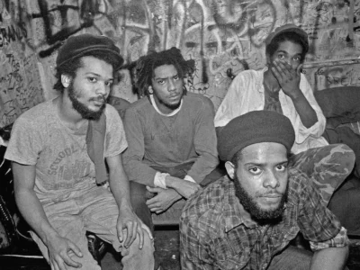 Black-and-white photograph of four Bad Brains group seated closely together in a cramped, graffiti-covered backstage or club dressing room. They face the camera with serious, introspective expressions—one leaning forward in the foreground wearing a cap and plaid shirt, another with short dreadlocks centered, while the others sit to the side, one partially covering his mouth. The walls are densely layered with handwritten tags and drawings, creating a raw, gritty atmosphere that suggests underground music culture and an intimate, unpolished moment behind the scenes