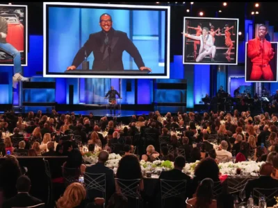 A large formal awards ceremony unfolds in a grand ballroom, where a Eddie Murphy stands at a podium on a brightly lit stage. Behind him, oversized screens display his image alongside framed visuals of past performances, adding context and celebration to the moment. The audience, dressed in formal attire, fills round banquet tables adorned with floral centerpieces, all facing the stage. Blue-toned lighting and a live band setup to the side enhance the atmosphere, emphasizing the scale and prestige of the event