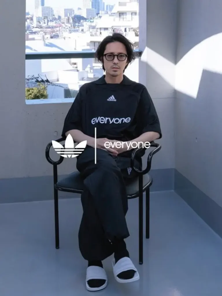 A seated portrait of a model wearing a black Adidas “everyone” outfit, positioned on a simple chair in a sunlit balcony setting. The individual wears a loose black short-sleeve shirt with a small Adidas logo at the chest and “everyone” printed across the front, paired with relaxed black pants. They sit with legs crossed, hands resting calmly, and wear round eyeglasses and black slide sandals with white soles. Behind them, a cityscape is visible through the open balcony, adding depth to the otherwise minimal scene. Overlaid in the foreground are the Adidas trefoil logo and the word “everyone,” reinforcing the collaborative branding