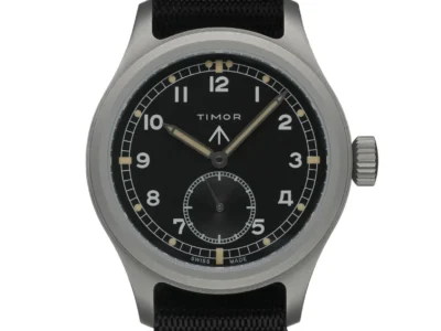 A clean studio shot of a brushed metal field watch with a black dial, luminous Arabic numerals, and a small seconds subdial, paired with a black fabric strap against a plain light background