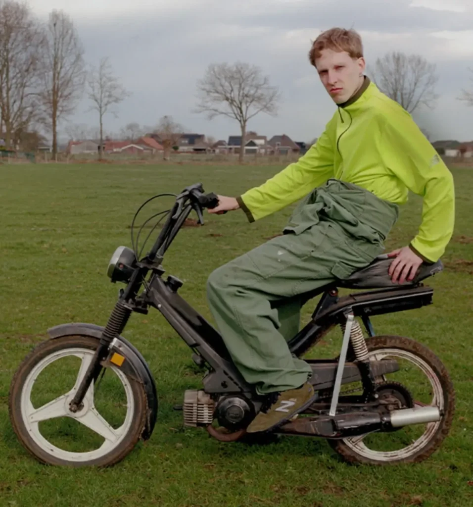 Young man in lime green jacket sitting on a black moped in a grassy field under cloudy skies