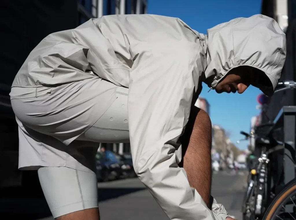 A man is bent forward on a city street, adjusting something near his shoes—likely laces—while wearing a minimalist, monochrome outfit. He has on a light beige hooded windbreaker with elasticated openings that cinch tightly around his head and wrists, paired with matching fitted shorts or compression-style bottoms in a similar tone. The fabric appears technical and lightweight, suggesting performance or athleisure wear