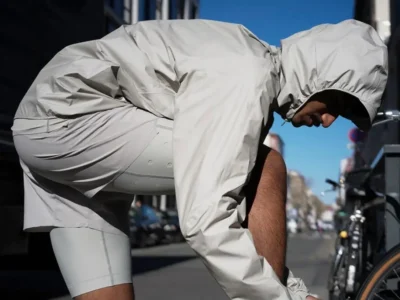A man is bent forward on a city street, adjusting something near his shoes—likely laces—while wearing a minimalist, monochrome outfit. He has on a light beige hooded windbreaker with elasticated openings that cinch tightly around his head and wrists, paired with matching fitted shorts or compression-style bottoms in a similar tone. The fabric appears technical and lightweight, suggesting performance or athleisure wear