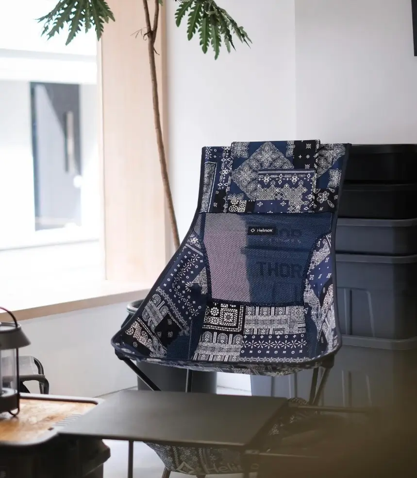 Compact indoor setup featuring a patterned Helinox camping chair with navy and white bandana-style patchwork fabric, positioned beside a small black folding table, with a lantern on a tray in the foreground and a tall indoor plant by a window adding soft natural light