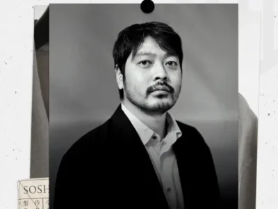 Black-and-white portrait of Soshi Otsuki in a tailored jacket and open-collar shirt, gazing directly with a composed, restrained expression against a minimal studio backdrop