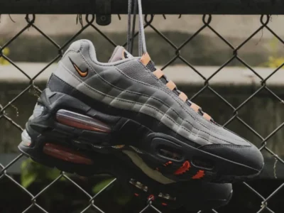 A pair of Nike Air Max 95 sneakers in layered grey and black tones with orange accents, suspended by their laces against a chain-link fence, showcasing the sculpted upper, mini Swoosh detail, and visible Air cushioning units with a slightly worn, urban backdrop