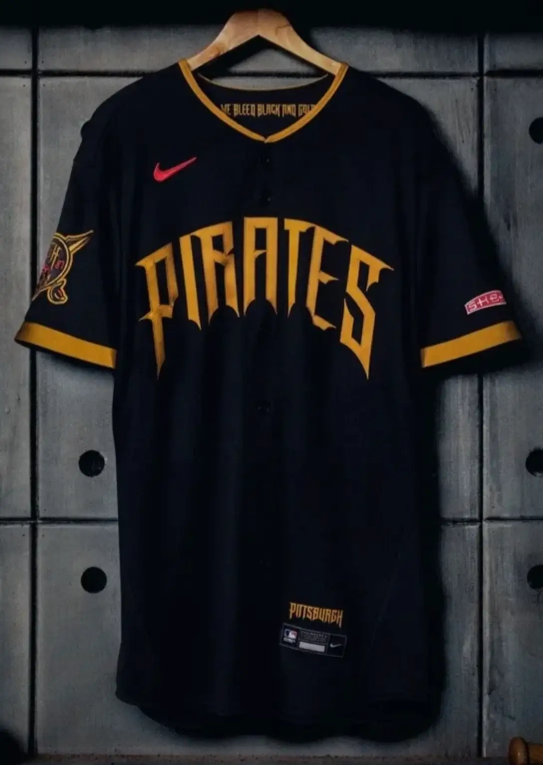 Front view of a black Pittsburgh Pirates baseball jersey hanging on a wooden hanger against a locker-style backdrop. The jersey features bold gold “PIRATES” lettering across the chest in a gothic-style font, a red Nike Swoosh on the upper chest, gold trim along the collar and sleeves, and a team patch on the sleeve, with “PITTSBURGH” labeling near the lower front